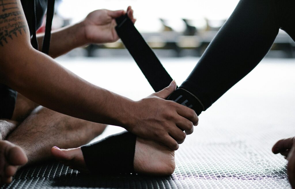 Close-up of hands tying a black strap around a partner's ankle on a gym mat, likely securing a fitness or martial arts band
