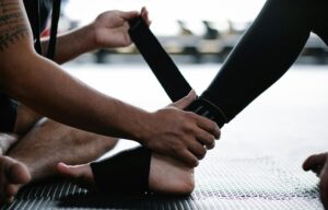 Close-up of hands tying a black strap around a partner's ankle on a gym mat, likely securing a fitness or martial arts band