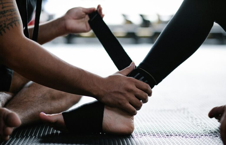 Close-up of hands tying a black strap around a partner's ankle on a gym mat, likely securing a fitness or martial arts band