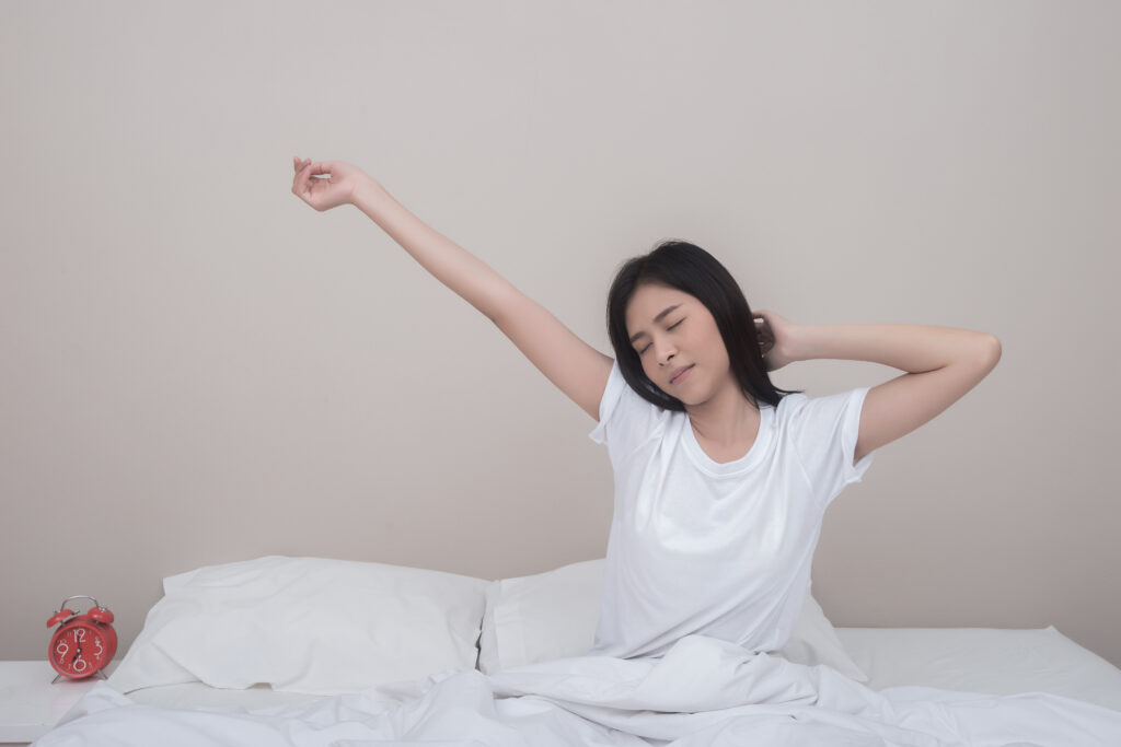 Woman waking up in bed, stretching with eyes closed; red alarm clock on nightstand to the left.