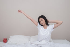 Woman waking up in bed, stretching with eyes closed; red alarm clock on nightstand to the left.