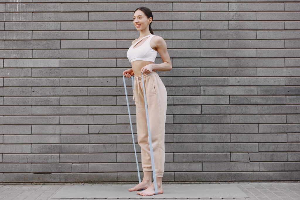 Woman in a white sports bra and beige sweatpants stands barefoot against a brick wall, holding light blue resistance bands for exercise.